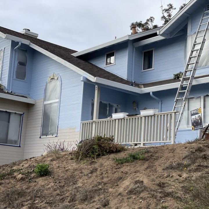 A blue house with multiple ladders and protective covering on windows, depicting an exterior painting project in progress.