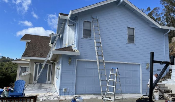 A light blue house with ladders and protective sheets on the windows, showcasing preparation for an exterior painting job.